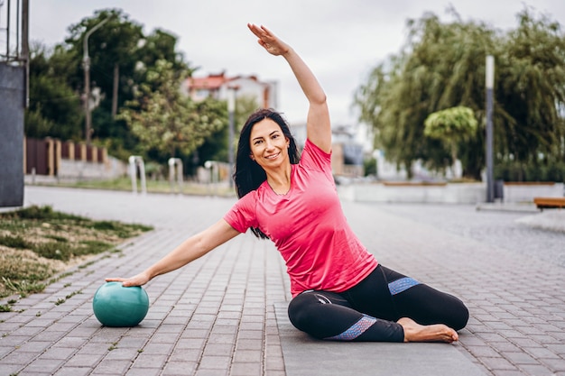 Sportive jeune femme aux cheveux longs, faire des exercices d'étirement avec un ballon spécial dans la rue. Mode de vie actif, concept de yoga