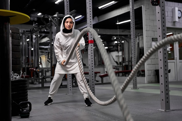 Sportive musulmane en forme et tonique travaillant dans une salle de sport d'entraînement fonctionnel faisant des exercices de crossfit avec des cordes de combat, portant un hijab sportif. motivation d'entraînement cross-fit