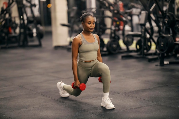 Une sportive noire en forme faisant des sauts avec des haltères au gymnase.