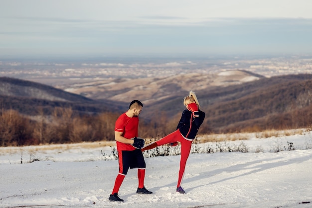 Sportive s'entraînant et donnant des coups de pied dans la nature lors d'une journée d'hiver enneigée avec son entraîneur. Boxe, fitness d'hiver, fitness en plein air