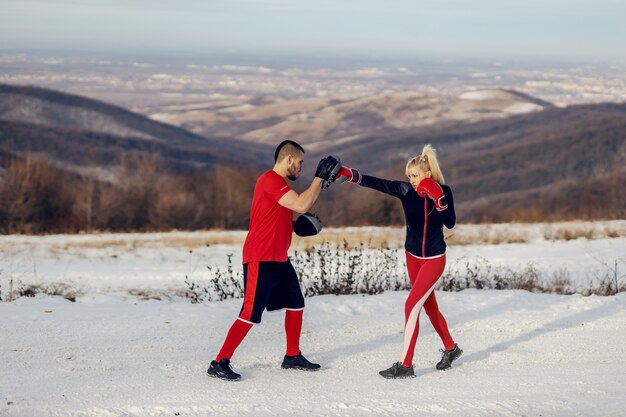 Sportive s'entraînant avec des gants de boxe dans la nature au jour d'hiver enneigé avec son entraîneur. Boxe, fitness d'hiver, fitness en plein air