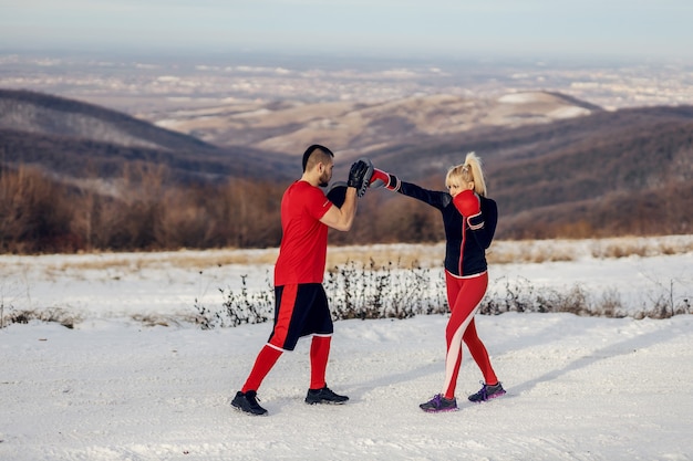 Sportive s'entraînant avec des gants de boxe dans la nature lors d'une journée d'hiver enneigée avec son entraîneur. Boxe, fitness d'hiver, fitness en plein air