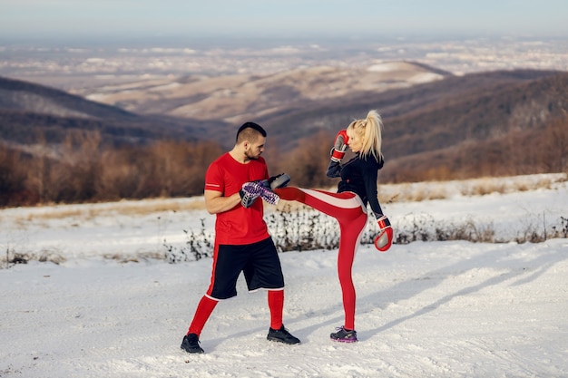 Sportive s'entraînant avec des gants de boxe dans la nature lors d'une journée d'hiver enneigée avec son instructeur. Elle donne des coups de pied. Boxe, fitness d'hiver, fitness en plein air