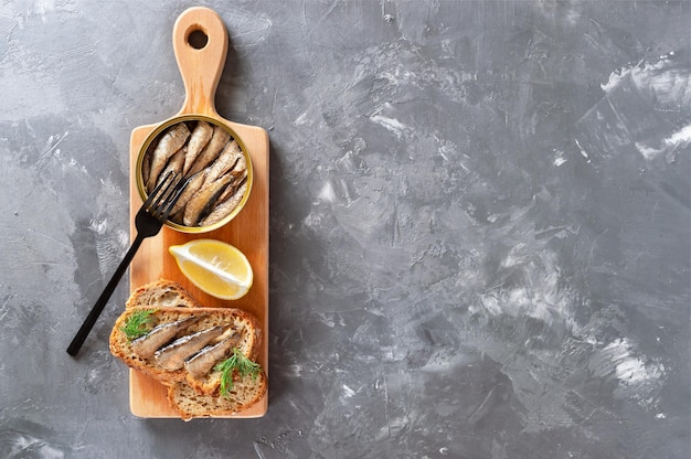 Sprat fumé à l'huile dans une boîte de conserve ouverte et des sandwichs au poisson sur une planche de bois sur fond de béton avec espace de copie. Vue de dessus, mise à plat