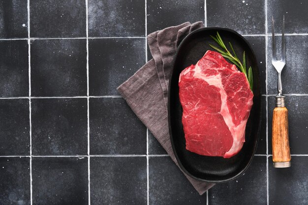 Steak de boeuf Bifteck de faux-filet de boeuf cru frais avec fourchette romarin sel et poivre sur une poêle à griller sur fond de pierre gris foncé ou noir Idée de publicité de boutique Vue de dessus Maquette pour idée de design