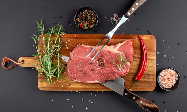 Steak de boeuf cru cuisson avec couteau et fourchette pour la viande et le romarin frais et les épices sur une planche à découper en bois fond noir