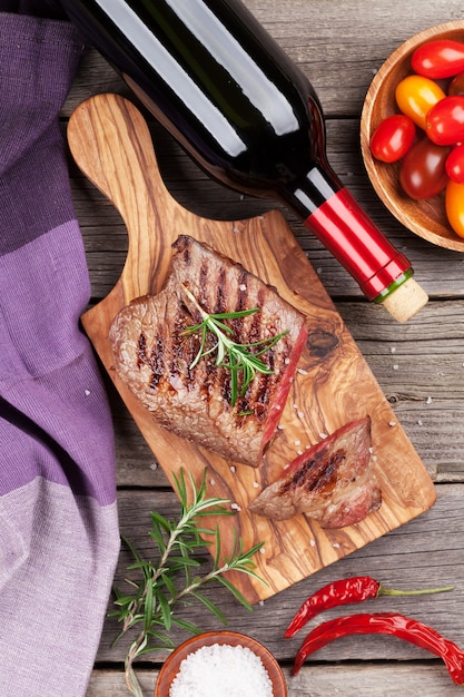 Steak de boeuf grillé au romarin, sel et poivre et bouteille de vin sur table en bois. Vue de dessus