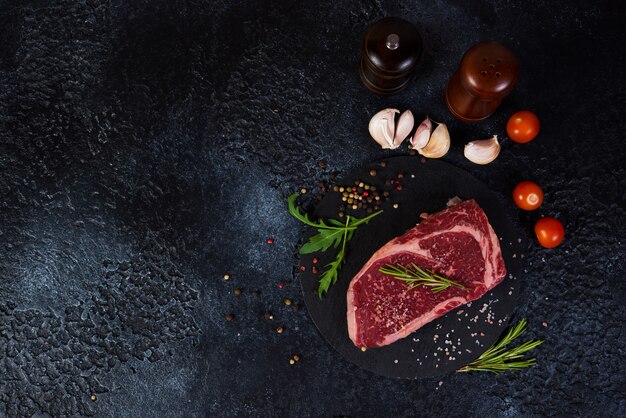 Un steak de bœuf marbré frais et cru se trouve sur un stand de charbon de bois à droite avec des épices, du sel et des herbes. Vue de dessus avec espace copie