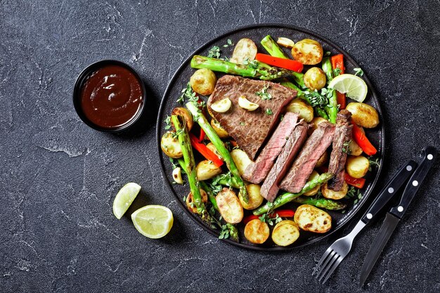 steak de boeuf avec pommes de terre nouvelles rôties, asperges et poivron rouge sur une assiette noire avec sauce barbecue sur une table en béton, plat, espace libre