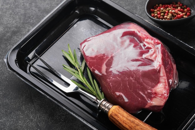 Steak de boeuf Steak de boeuf cru frais avec fourchette et romarin dans un emballage en plastique noir sous vide sur fond de pierre noire Emballage Vacoom Vue de dessus Maquette pour idée de conception