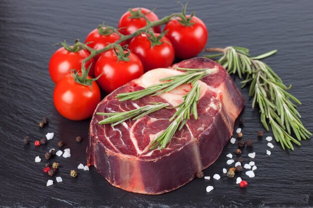 Steak de boeuf à la viande crue fraîche avec os avec épices, romarin, tomates et ail sur la surface de l'ardoise noire. ingrédients de cuisson crus