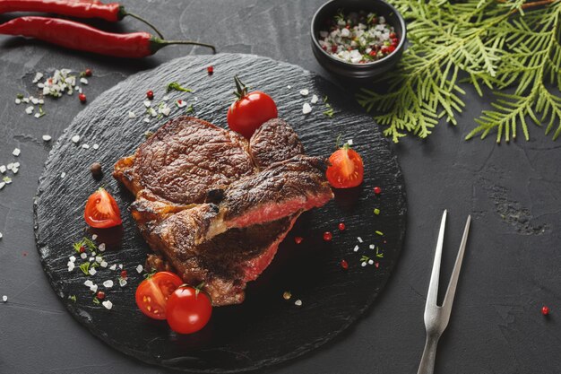 Steak de faux-filet sur plaque sombre avec des brindilles de thuya décorées de tomates, piment, épices et fourchette à viande, gros plan