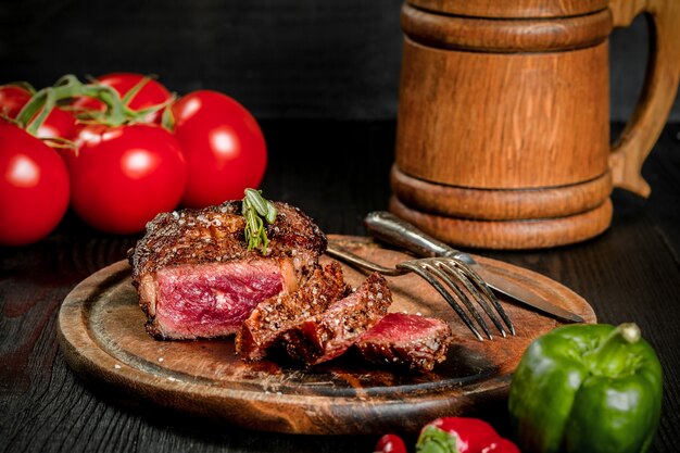 Steak grillé assaisonné d'épices et d'herbes fraîches servi sur une planche en bois avec chope de bière en bois, tomate fraîche, poivrons rouges et verts. Fond en bois noir. Nature morte