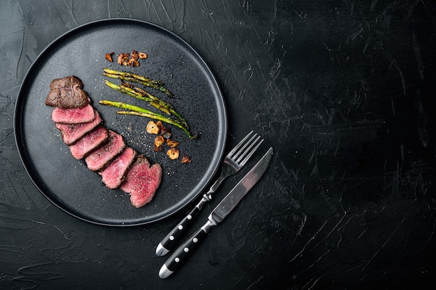 Steak juteux de bœuf saignant moyen Filet Mignon ou Filet d'oeil, avec oignon et asperges, sur assiette, avec couteau et fourchette à viande, sur pierre noire