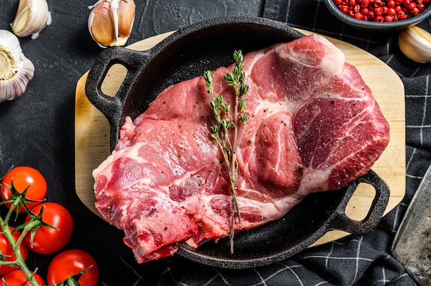 Steak de porc cru dans une poêle. Morceau de viande crue prête à être préparée avec des légumes verts et des épices. Fond noir. .
