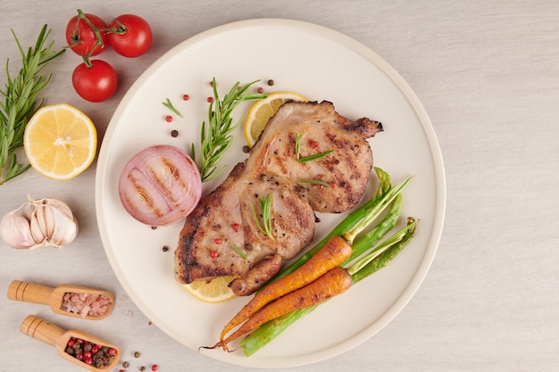 Photo steak de porc grillé d'un barbecue d'été servi avec légumes, asperges, carottes miniatures, tomates fraîches et épices. steak grillé sur plaque blanche sur surface en pierre. vue de dessus.