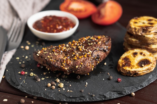 Steak de porc rôti sur planche de pierre noire sur fond sombre avec sauce tomate et pommes de terre frites