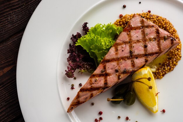 Steak de thon frit sur assiette blanche ronde avec assaisonnement et salade verte au citron et câpres sur table en bois