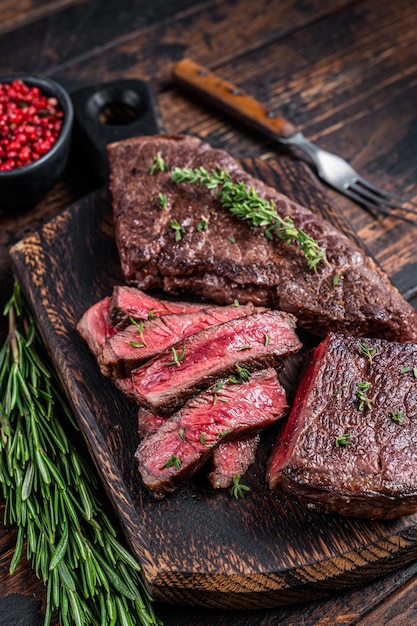 Steak de viande de boeuf en tranches grillées sur une planche à découper avec des herbes. Fond en bois sombre. Vue de dessus.