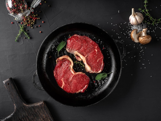 Steaks crus avec assaisonnements dans une poêle en fer fond noir Viande fraîche herbes ail champignon poivre sel planche de bois Gros plan Vue de dessus