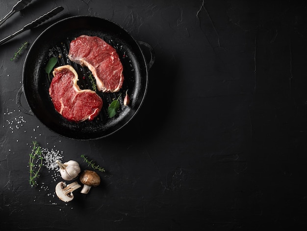 Steaks crus avec assaisonnements dans une poêle à griller en fer fond noir Viande fraîche herbes ail poivre sel champignon pinces Gros plan Vue de dessus