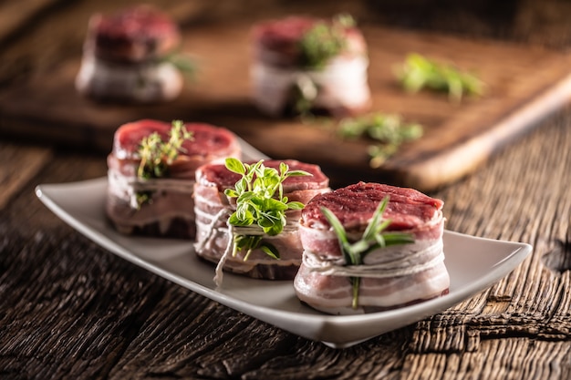 Steaks crus enveloppés de bacon et d'herbes fraîches sur une assiette et une surface en bois vintage.