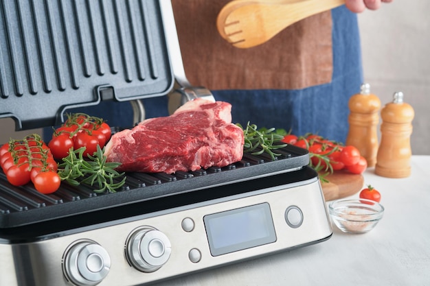 Steaks crus de première qualité sur un gril électrique Main masculine du chef tenant un steak Ribeye sur un gril électrique moderne avec tomate cerise au romarin et poivre Processus de cuisson dans une cuisine moderne Espace de copie vue de dessus
