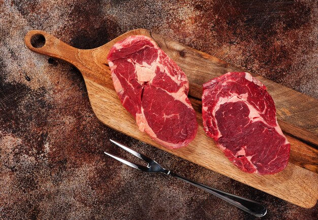 Steaks de faux-filet crus sur une planche à découper en bois et une fourchette à viande sur fond de béton brun, vue de dessus