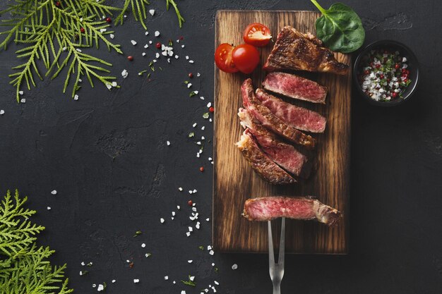 Steaks de faux-filet rares aux herbes et épices avec fourchette à viande et tomate sur fond sombre, décorés de brindilles de thuya, espace pour copie, vue de dessus