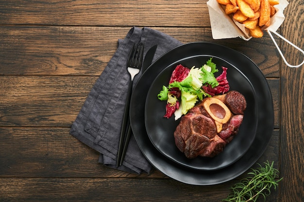 Steaks Osso Buco Tranches de boeuf grillé Osso Buco ou bifteck de viande de portier avec garni de salade et frites sur planche de marbre noir sur fond de bois ancien Vue de dessus Maquette