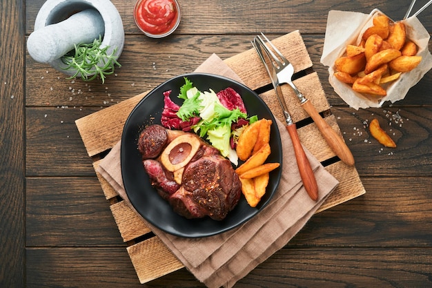 Steaks Osso Buco Tranches de boeuf grillé Osso Buco ou bifteck de viande de portier avec garni de salade et frites sur planche de marbre noir sur fond de bois ancien