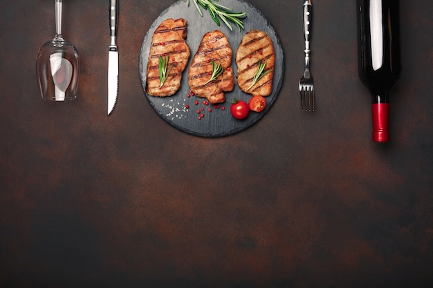Steaks de porc grillés sur pierre avec bouteille de vin, verre à vin, couteau et fourchette sur fond rouillé