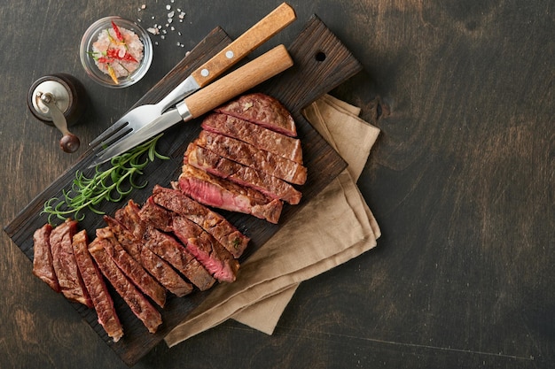 Steaks Tranches de steak de viande grillée New York ou Ribeye avec épices romarin et poivre sur planche de marbre noir sur fond de bois ancien Vue de dessus Maquette