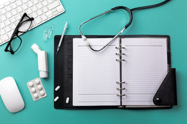 Stéthoscope dans le bureau des médecins avec ordinateur portable, clavier, souris, lunettes, seringue, ampoules, inhalateur et pilules. Vue de dessus avec place pour votre texte.