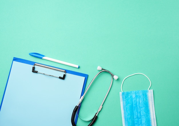 Stéthoscope en métal, porte-papier avec une feuille bleue vierge, masque médical bleu jetable sur fond vert