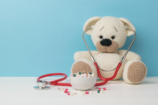 Un stéthoscope rouge sur un ours en tricot blanc et des pilules dans un bol.