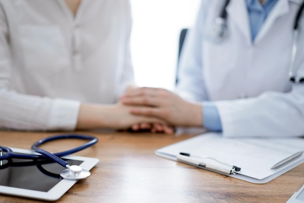 Le stéthoscope et la tablette sont allongés sur la table en bois tandis que les mains du médecin rassurent un patient à l'arrière-plan. Notion de médecine