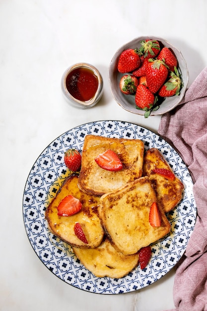 Stock de pain doré avec des fraises fraîches sur une assiette en céramique, du sirop d'érable dans une cruche en céramique et une serviette en tissu rose sur une surface blanche. Vue de dessus, pose à plat. Copier l'espace