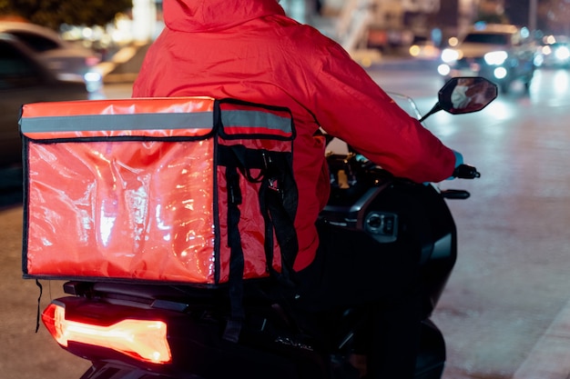 Stock photo d'un livreur de nourriture en uniforme rouge portant une boîte de livraison de nourriture à livrer au client pour la commande pendant la pandémie de COVID-19 et le verrouillage dans la ville la nuit en Thaïlande.