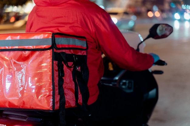 Stock photo d'un livreur de nourriture en uniforme rouge portant une boîte de livraison de nourriture à livrer au client pour la commande pendant la pandémie de COVID-19 et le verrouillage dans la ville la nuit en Thaïlande.