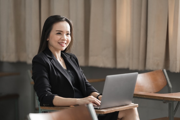 Stock photo portrait d'une enseignante asiatique joyeuse et confiante dans un uniforme de costume noir avec une tablette numérique et un ordinateur portable pour enseigner la langue moderne en classe