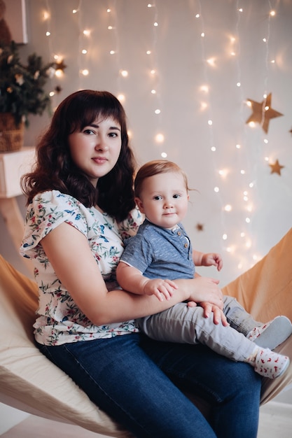Stock photo portrait de mignon petit garçon avec sa mère assis sur la balançoire contre la guirlande lumineuse sur le mur en arrière-plan. Mère embrassant son fils et regardant la caméra. Concept de Noël.