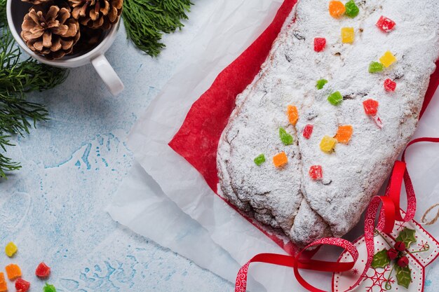 Stollen à grains entiers avec raisins secs et sucre en poudre sur une serviette en lin avec un tamis, ruban rouge sur le béton enneigé bleu clair