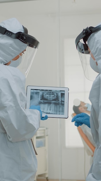 Stomatologues avec des combinaisons de ppe tenant une tablette numérique avec des dents x ray analysant les soins dentaires pour l'opération d'implant à la clinique médicale. Dentistes utilisant la technologie de numérisation pendant la pandémie