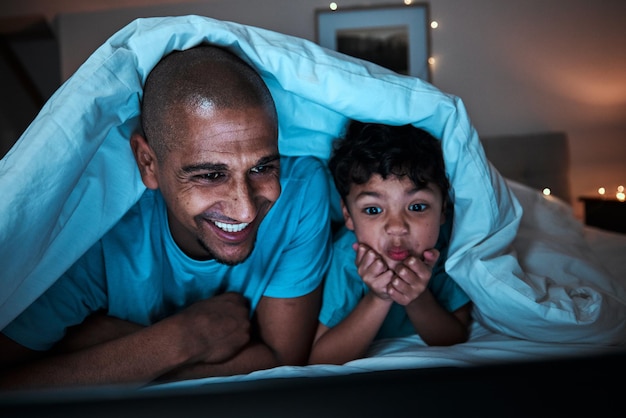 Streaming nocturne et père avec enfant dans un lit pour un fichier de film ou un divertissement par abonnement dans leur maison Regarder la télévision et parent avec un garçon enfant dans une chambre à coucher heureux et s'amusant