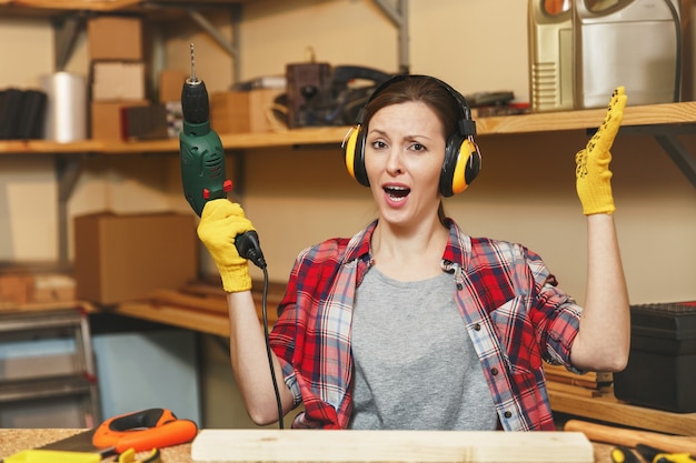 Le stress en colère a bouleversé une jeune femme aux cheveux bruns en chemise à carreaux, des écouteurs insonorisés travaillant dans un atelier de menuiserie à la table, perçant avec des trous de perceuse électrique dans un morceau de bois, fabriquant des meubles.