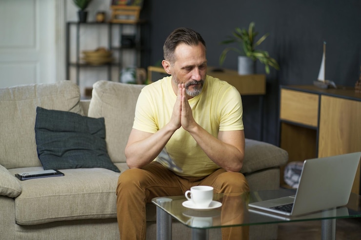 Photo stress émotionnel homme mûr assis sur un canapé avec les mains réunies à côté d'une table basse avec un ordinateur portable et une tasse dessus à l'intérieur de la maison portant une crise d'âge moyen décontractée dévore un homme solitaire perplexe