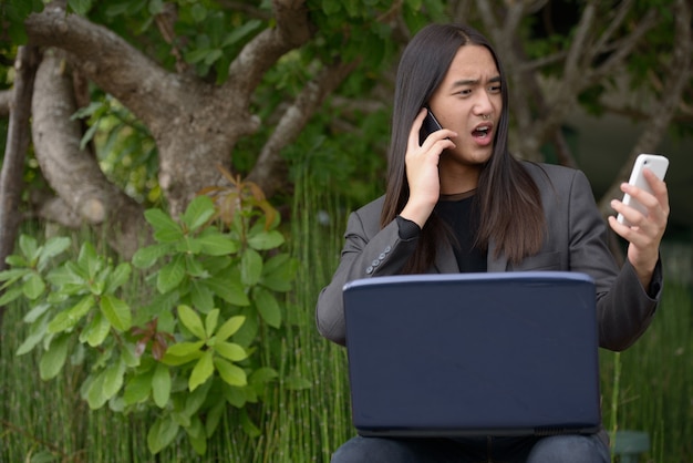 Stressé jeune homme d'affaires asiatique aux cheveux longs à l'aide d'un ordinateur portable et de deux téléphones tout en travaillant dans le parc