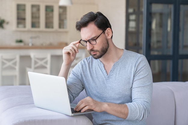 Stressé triste fatigué sérieux jeune homme caucasien homme d'affaires étudiant professeur travaillant à l'aide d'un ordinateur portable assis sur le canapé à la maison à l'intérieur problèmes d'affaires de bureau à la maison