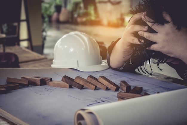 Stressed Engineering est tombé avec un plan et un casque. Un jeune homme est assis à son bureau et tient ses mains sur sa tête.